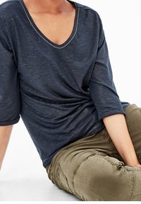 Navy blue, lightweight V-neck shirt with three-quarter sleeves, featuring a draped design and subtle sheen. Paired with olive green pants.