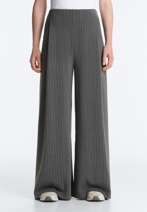 Személy széles szárú, szürke, vékony csíkos nadrágot és fehér tornacipőt visel, egyszerű fehér háttér előtt áll.