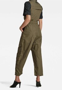 Kvinna i olivgrön cargojumpsuit med korta ärmar och svarta högklackade mules, stående med händerna i fickorna vänd bort.