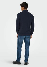 Pull en motif marine avec un col haut, associé à un jean bleu clair et des bottes noires, vu de dos. La texture est côtelée et en zigzag.