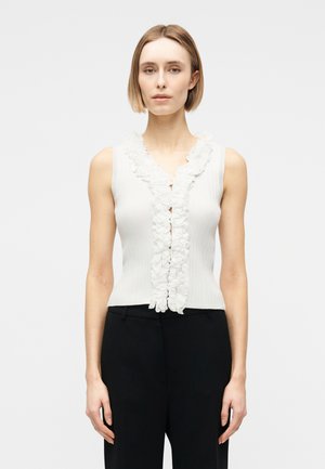 Kvinne med kort blondt hår iført ermeløs hvit topp med volangdetaljer foran og svarte bukser, stående mot en ensfarget lys bakgrunn.