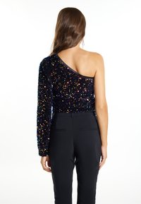 Top nero in paillettes con una sola spalla, caratterizzato da una silhouette aderente e una texture scintillante, abbinato a pantaloni neri su misura.