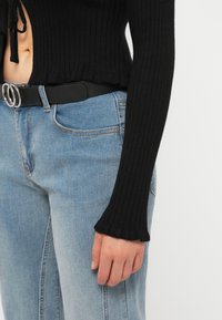 Femme portant un jean bleu clair avec une ceinture noire et un haut noir à manches longues côtelé, à ourlet court et avec un détail noué en tissu.