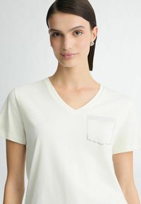 V-hals, kortærmet t-shirt i lys beige cotton med en tekstureret lomme detalje, der har et subtilt grafisk præg. Glat, åndbart stof.