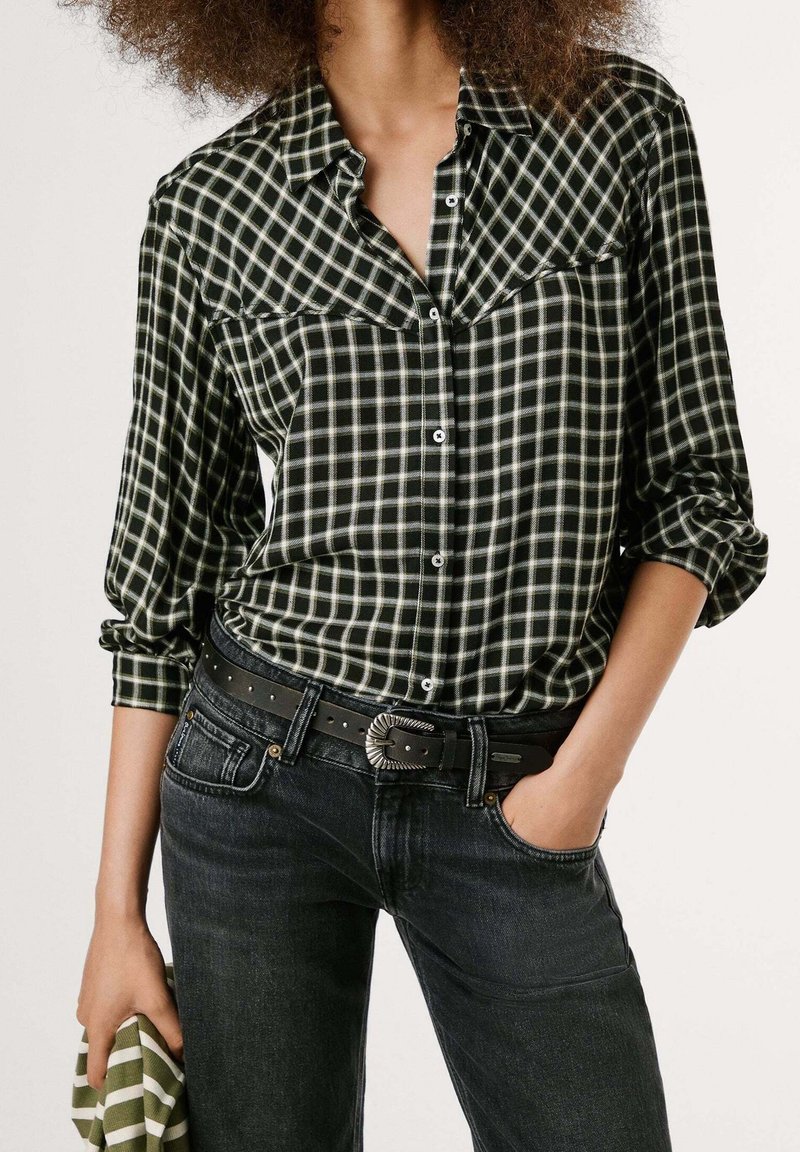 Personne portant une chemise à manches longues à carreaux noirs et blancs rentrée dans un jean noir, tenant un tissu rayé dans une main.