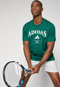 T-shirt de algodão verde com impressão em creme "ADIDAS TENNIS CLUB". O modelo segura uma raquete de ténis preta com detalhes em azul, vestindo calções brancos.