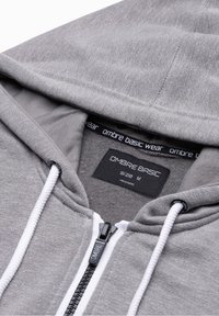 Sweat à capuche gris à fermeture éclair, avec une texture douce, une étiquette noire et blanche à l'intérieur, des cordons fins blancs et une fermeture éclair métallique. Taille M.