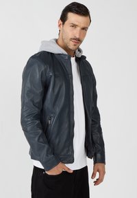 Dunkelblaue Lederjacke mit taillierter Passform, die mit einer grauen Kapuze, die aus dem Kragen hervorlugt, und seitlichen Reißverschlusstaschen ausgestattet ist.