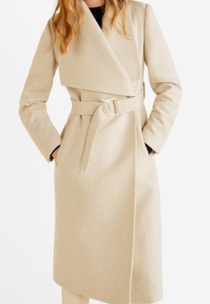 Abrigo largo beige con cinturón y solapas anchas, llevado sobre una camiseta negra, con las manos del modelo metidas en los bolsillos, recortado por debajo de los hombros.