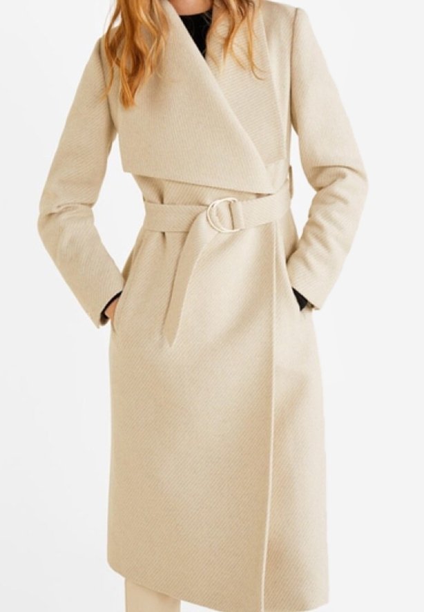 Long manteau beige ceinturé avec de larges revers porté par-dessus un haut noir, les mains du mannequin dans les poches, cadré en dessous des épaules.