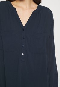Donna che indossa una blusa blu navy scuro con maniche lunghe, due tasche sul petto e quattro bottoni lungo la parte anteriore.