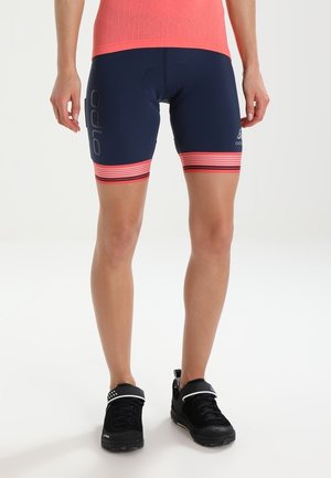 Persoon die marineblauwe en koraalkleurige fietsshorts met gestreepte zomen draagt, staand in zwarte sportschoenen tegen een witte achtergrond.