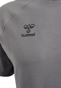 Graues T-Shirt mit glatter Textur, mit einem dunklen Logo im linken Brustbereich. Runder Halsausschnitt und kurze Ärmel.