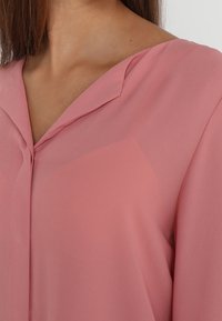 Blouse rose clair avec un col en V et des manches longues, réalisée dans un matériau transparent et fluide. Présente une légère brillance et une texture douce.