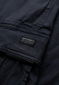 Gros plan sur un tissu noir avec des coutures détaillées et une petite étiquette rectangulaire de la marque Superdry cousue sur le matériau.