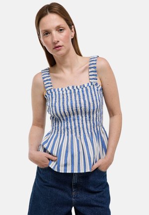 Femme portant un haut sans manches rayé bleu et blanc à larges bretelles et un jean bleu foncé, debout les mains dans les poches.
