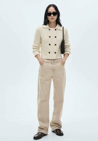 Cardigan lavorato a maglia color crema con bottoni neri e colletto a camicia, abbinato a pantaloni beige a gamba larga. Il modello indossa occhiali da sole neri e tiene una borsa piccola.