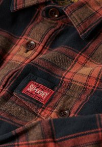 Punainen ja musta ruudullinen flanellipaita, jossa on kudottu rakenne, ruskeat napit ja punainen Superdry -etiketti rinnustaskussa.