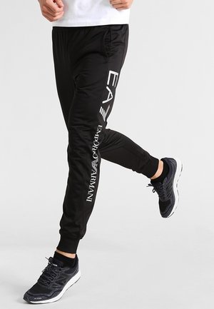 Persona vistiendo pantalones deportivos negros Emporio Armani EA7 y zapatos deportivos negros, de pie con una pierna levantada y doblada.