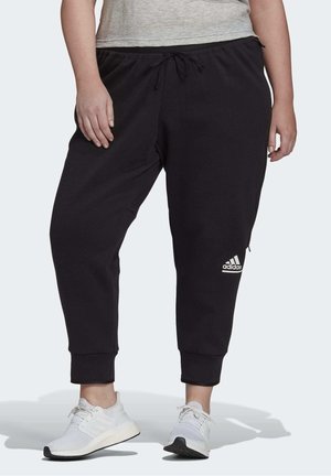 Person wearing black Adidas joggers and white athletic shoes, standing against a plain light background.