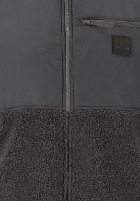 Musta fleece-tyylinen vetskari-liivi, jossa on sileä yläosa ja teksturoitu alaosa. Varustettu vetoketjullisella rintataskulla ja logopatchilla.