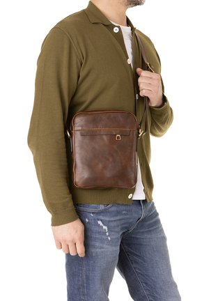 Man in olijfgroene cardigan en blauwe spijkerbroek met een bruine leren crossbodytas met een gesp detail.