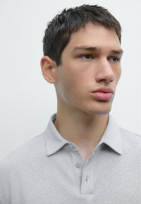 Svetle šedé polo tričko s límečkem a dvěma knoflíky, vyrobené z měkké, strukturované tkaniny. Modelova tvář je viditelná, má krátké vlasy.