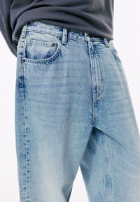 Persoon in lichtblauwe hoogwaisted denim jeans en een donkergrijze, in de broek gestopte, lange mouwen shirt, dat tot halverwege de bovenbenen komt.