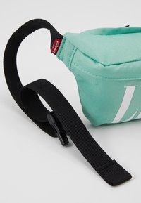 Marsupio in tessuto azzurro chiaro di forma rettangolare, con un grande logo bianco e una cinghia nera regolabile con clip.