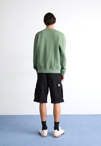 Grüner Sweatshirt mit Rundhalsausschnitt, schwarze Cargo-Shorts und weiße Sneaker; Merkmale umfassen einen gerippten Saum und einen Aufnäher auf den Shorts.