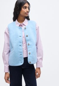 Ανοιχτό μπλε γιλέκο από φλις με στρογγυλή λαιμόκοψη, διαθέτει έξι κουμπιά και πλαϊνές τσέπες, φορεμένο πάνω από ένα ροζ ριγέ πουκάμισο.