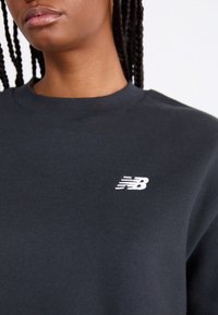Sweatshirt noir avec col ras du cou côtelé et logo 'NB' brodé en blanc. Le tissu semble doux avec une texture lisse.