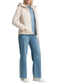 Veste matelassée beige légère avec capuche, fermeture éclair et cordons, portée sur une chemise en denim et associée à un jean large bleu et des baskets blanches.
