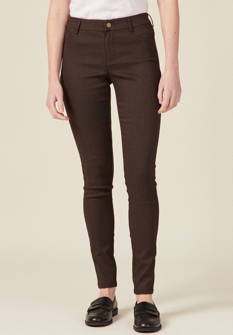 Pantalon slim-fit marron à carreaux avec poches avant et fermeture à bouton, porté avec des mocassins noirs par une personne se tenant devant un fond clair.