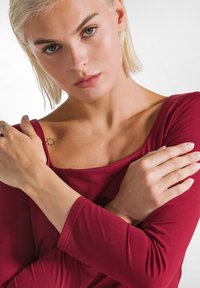 Bordeauxrode top met lange mouwen, een brede halslijn, een gladde textuur en een aansluitend ontwerp, waarbij een schoudertatoeage zichtbaar is. Geen verdere details aanwezig.
