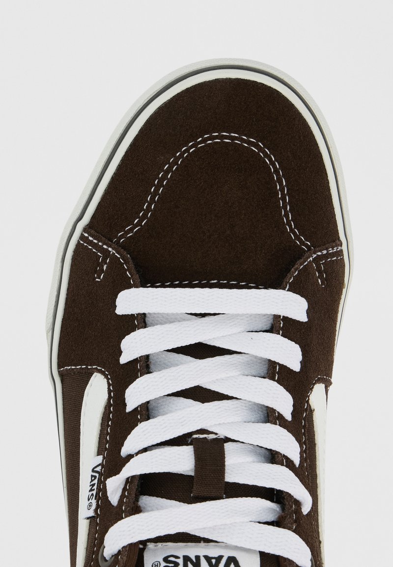 Primer plano de una zapatilla Vans de ante marrón con cordones blancos y costuras blancas contrastantes sobre un fondo liso.