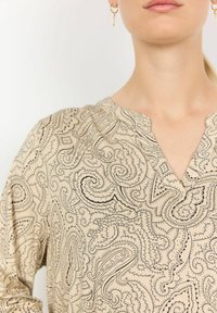 Beżowa bluzka z czarnym wzorem paisley, z dekoltem w lekki kształt V i długimi rękawami. Miękka tekstura materiału i luźny krój.