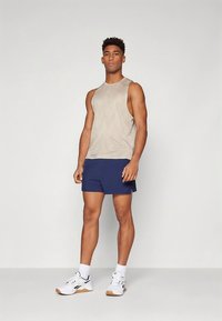 Világos bézs színű ujjatlan trikó és tengerészkék rövidnadrág. A trikón enyhe minta található, a rövidnadrág sima anyagból készült. A sportcipő teszi teljessé az öltözéket.