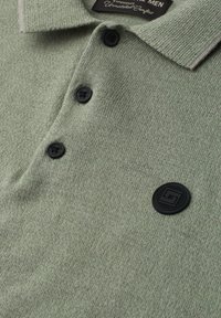 Polo en maille vert clair avec trois boutons noirs et un logo circulaire noir sur la poitrine, montrant le col et une partie de l’étiquette.