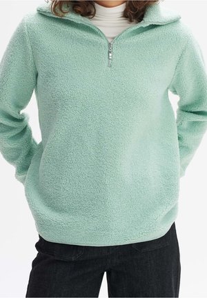 Personne portant un pull polaire vert clair avec une demi-fermeture éclair sur un col roulé blanc et un pantalon noir, mains derrière le dos.