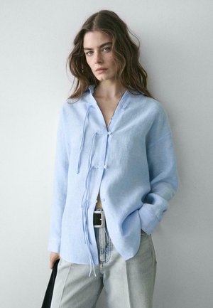 Femme aux cheveux bruns ondulés portant une chemise bleu clair à manches longues avec des nouettes à l'avant, un jean gris et une ceinture noire, debout contre un mur uni.