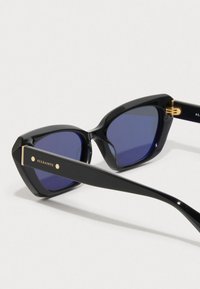 Sorte kattøye solbriller med blåtonede linser og gull «AllSaints»-merking på stengene, hvilende på en hvit overflate.