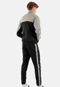 Sportanzug in Schwarz und Grau. Die Jacke hat einen hohen Kragen und ein Logo auf der Rückseite. Die Hose hat seitliche Streifen und elastische Bündchen. Glatte Textur.