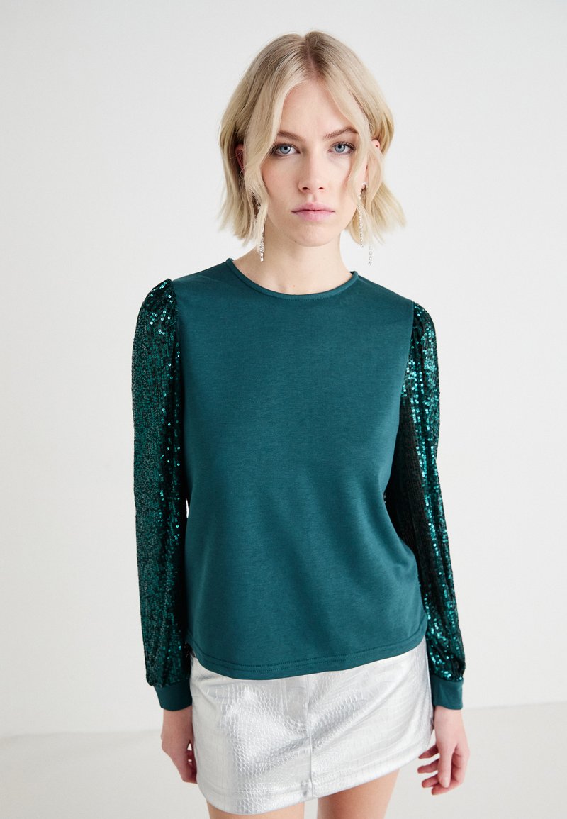 Top de manga larga color verde azulado con mangas acentuadas con lentejuelas, combinado con una falda plateada reflectante. Tejido suave con un brillo sutil y puños acanalados.