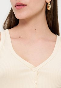 Gros plan d’une femme portant un haut sans manches côtelé crème et des boucles d’oreilles créoles dorées, montrant son cou et sa clavicule.