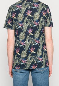 Bakvy av en person som bär en mörk skjorta med färgglad paradisfågel- och stora bladtryck, tillsammans med blå jeans.