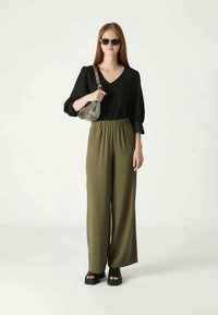 Woman with straight red hair wearing black V-neck blouse, olive wide-leg pants, black sandals, sunglasses, and carrying a small gray shoulder bag.