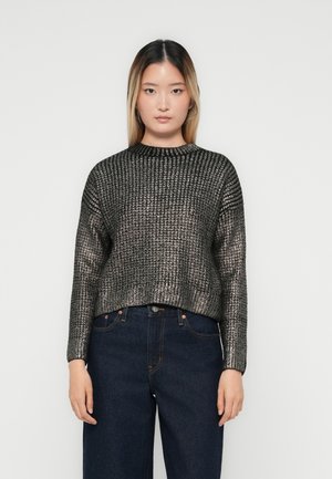 Ung kvinne iført en metallisk ribbet genser og mørkeblå jeans, stående mot en ensfarget hvit bakgrunn.