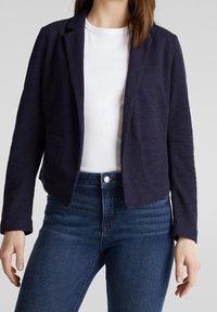 Blazer azul marino hecho de tela texturizada, con abertura frontal, dos bolsillos delanteros y una silueta entallada, combinado con una camisa blanca y jeans.