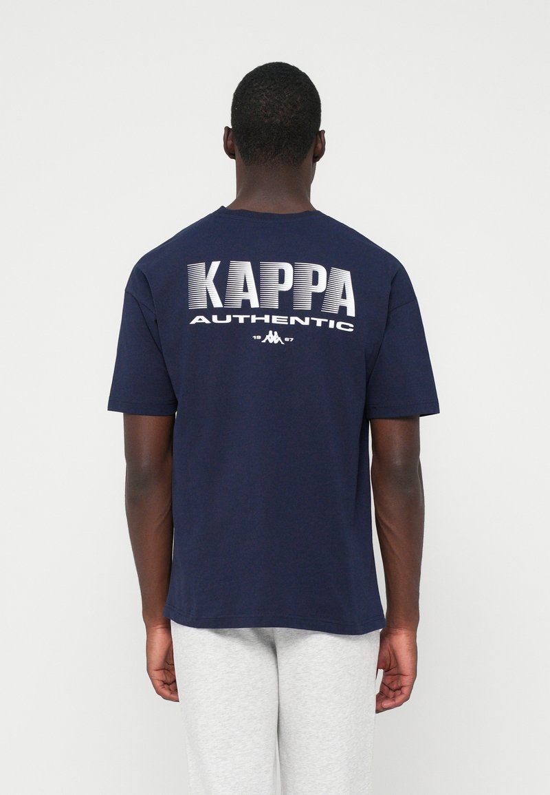 Persona con una camiseta Kappa Authentic azul marino con texto blanco en la parte posterior, combinada con pantalones gris claro, de pie de espaldas.